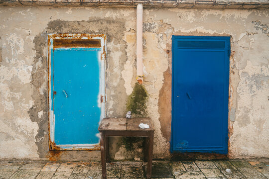 View of two weathered doors in light and dark blue on a textured, peeling wall with a small wooden table in between, located in Bari, Apulia, Italy.