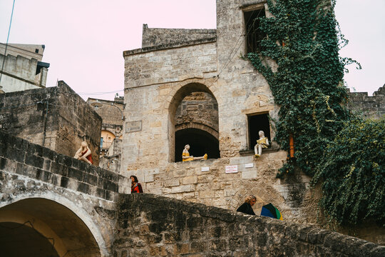 View of mannequins placed in the stone archways and windows of historic buildings in the Sassi di Matera, with people walking nearby in Matera, Basilicata, Italy.