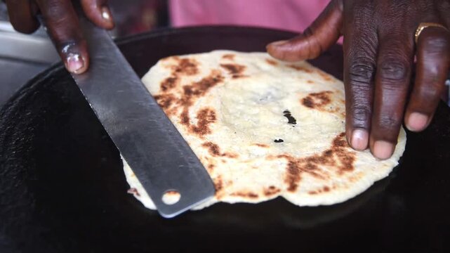 hands cooking flatbread roti on hot black griddle, toasted brown spots on dough and metal spatula, in close-up view, traditional street food preparation in slow motion