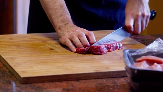 Dicing strips of stew beef into a large dice, with a chef's knife, in home kitchen, on a wooden cutting board, on a red granite tabletop. Close up