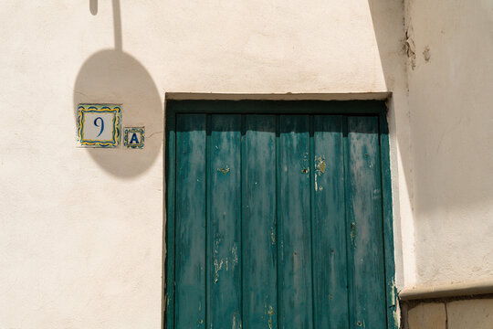 View of a weathered green wooden door with a ceramic house number tile on a light stucco wall in Capri, Campania, Italy.