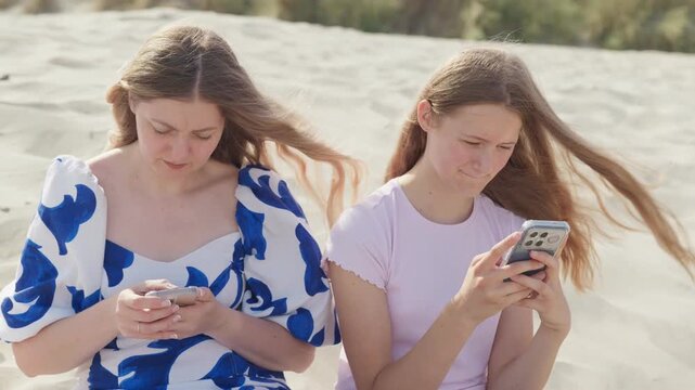 A woman and girl are sitting on the beach with phones the girl medium shot