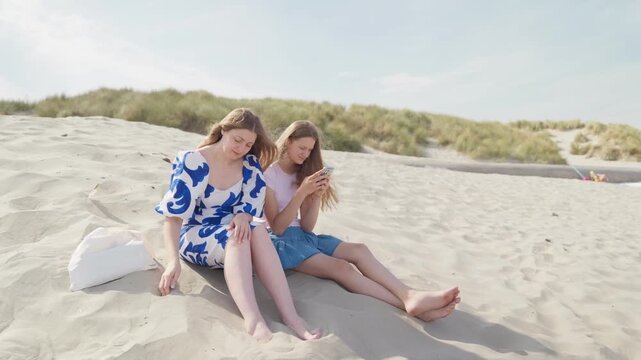 Woman scoops up sand in her hands and daughter is sitting with phone in hands, at the beach wide shot camera movement
