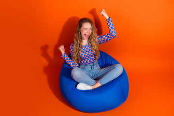 Naklejka na ściany i meble Cheerful young woman celebrating a moment of happiness while sitting on a vibrant blue bean bag against a bright orange background