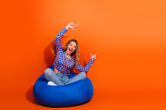 Charming woman sitting on a blue beanbag chair in front of a vivid orange background, energetically posing