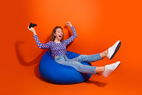 Excited young woman gaming with controller on beanbag against vibrant orange background, full of energy and joy