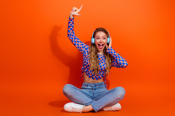Naklejka na ściany i meble Excited young girl wearing stylish outfit sitting cross-legged and enjoying music with headphones on bright orange background