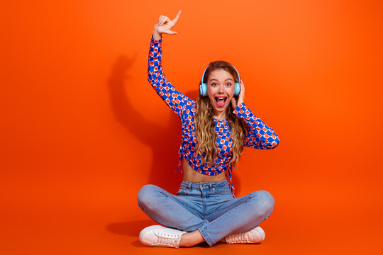 Excited young girl wearing stylish outfit sitting cross-legged and enjoying music with headphones on bright orange background