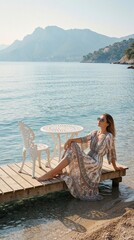 Fototapeta premium Stylish woman in long patterned dress sits on wooden dock by blue sea. Wears sunglasses, relaxes near white metal table, chair. Calm ocean water, distant mountains create peaceful summer vacation