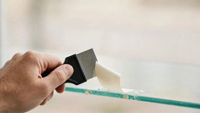 Hand using scraper to remove stubborn adhesive label from glass shelf
