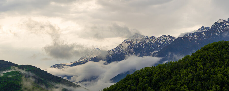 Panoramic view of Caucasus mountain ridge, snowy peaks and clouds over green forested hills