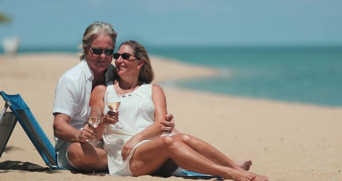 Senior couple relaxing on a tropical beach, enjoying a romantic moment with a kiss and glasses of wine, celebrating their retirement and love during a luxurious vacation on Samui island, Thailand
