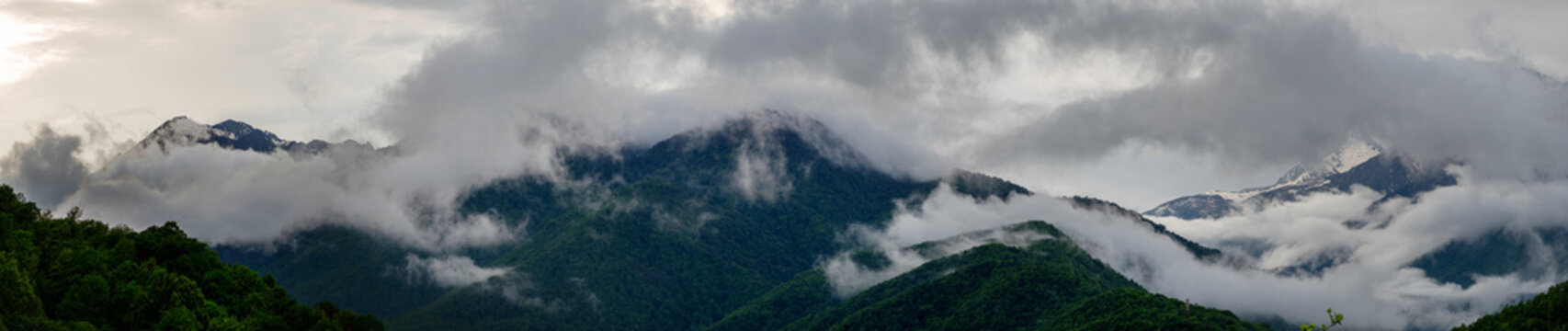 Ultra-wide panorama of misty green mountains and snowy peaks in low clouds, long banner