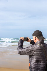A side view of a senior man wearing a cap and winter jacket taking a photo of the sea waves with a smartphone. Active retirement lifestyle, travel scenery. Travel theme. © Natalia