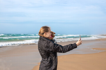 A young woman stands on a windy sandy coast recording a video of sea waves using a smartphone. Autumn or winter oceanside travel recreation layout. Travel theme. © Natalia
