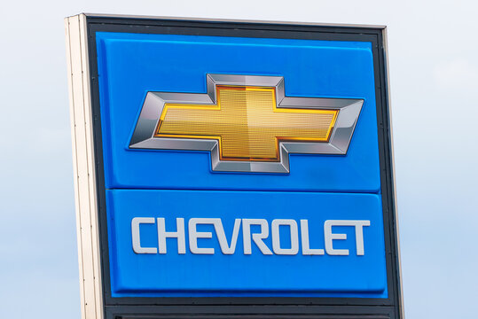 Chevrolet brand and sign in blue, white, silver and gold, with local and words. Taken at a dealership with light blue sky in the background.