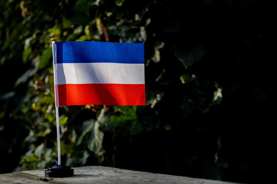Miniature Crimea regional flag rests on a slim pole over a weathered board with soft green foliage behind and gentle sunlight on the fabric offering a calm patriotic scene for study travel