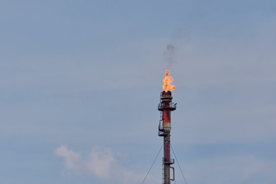 Gas flare stack chimney burning hydrocarbon gas with bright yellow flame against blue cloudy sky at petroleum refinery plant.