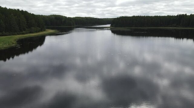 Drone flying low above a calm lake in a northern forest with reflection of grey clouds
