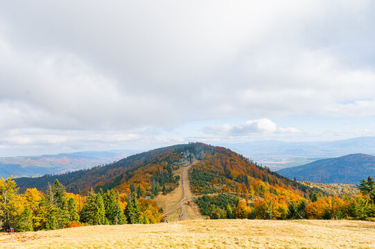 A wide panoramic view of a mountain ridge path winding toward a distant shelter surrounded by bright autumn foliage. Colorful orange and red trees cover the slopes under a soft cloudy sky.