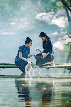 Side view of Two smiling young Thai women wearing traditional indigo blue dresses and patterned sinh skirts sitting on a bamboo bridge with a fishing net fishing, Sakon Nakhon, Isan region, Thailand