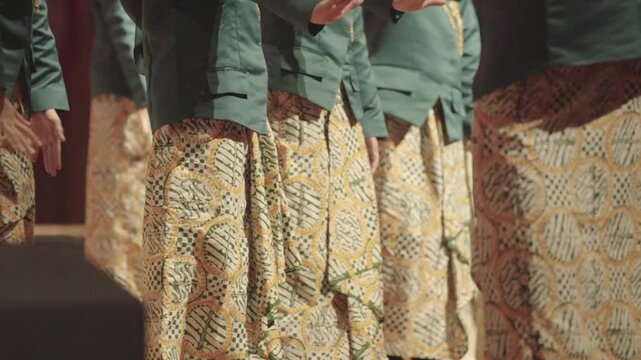 Close Up Panning Video of an Unrecognizable Young Asian Choir Group Standing in a Row on Stage During a Performance. Wearing Matching Green Jackets and Traditional Batik Patterned Skirts.