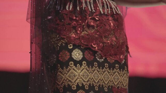 an Unrecognizable Asian Young Female Singer Gesturing on Stage During a Cultural Event. Wearing a Red Sequined Lace Top, Sheer Beaded Cape, and a Traditional Geometric Patterned Skirt Called Kebaya.