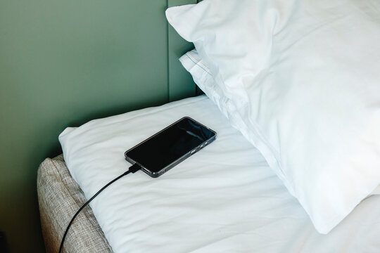 a smartphone charging on a neatly made bed beside two pillows