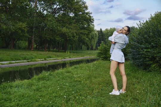 Woman stands on grass by canal in park while holding child. Trees and green plants surround them. Evening light casts soft shadows. They look happy and relaxed in outdoor setting