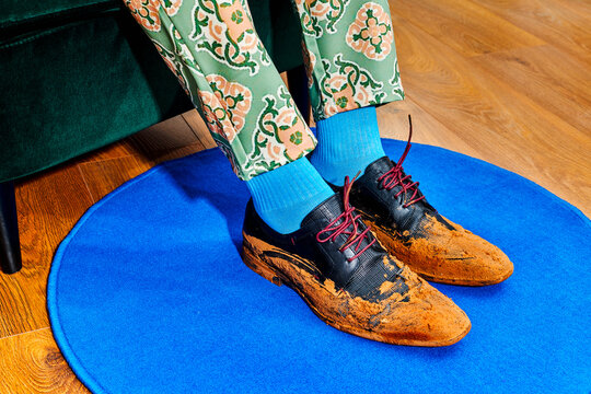 a man rests his feet in mud-covered black leather shoes on a blue rug