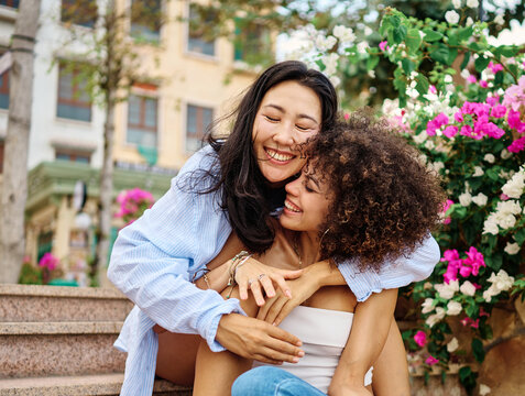 Two young women laugh and embrace on outdoor city steps, surrounded by bright flowering plants. Their joyful, affectionate moment captures friendship, intimacy and diverse connection in a casual urban