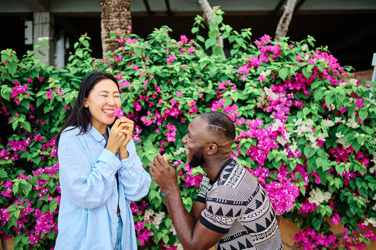 A laughing couple share a playful romantic moment by the seaside, surrounded by colorful bougainvillea and lush greenery. The man offers flowers while the woman smiles and looks upward, enjoying a sun