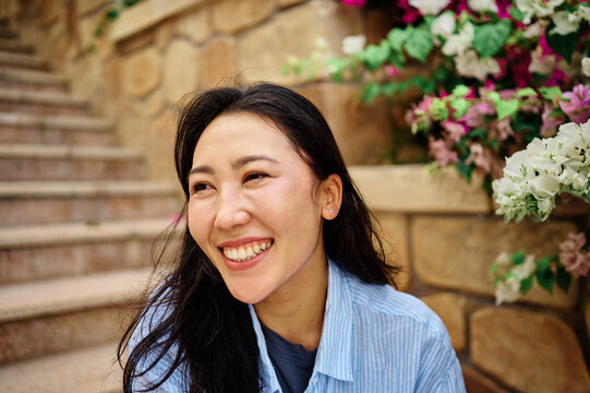 A relaxed young woman sits on stone steps beside blooming flowers, smiling with eyes closed in a candid outdoor portrait. Casual clothing and natural light create a joyful, peaceful mood, perfect for 