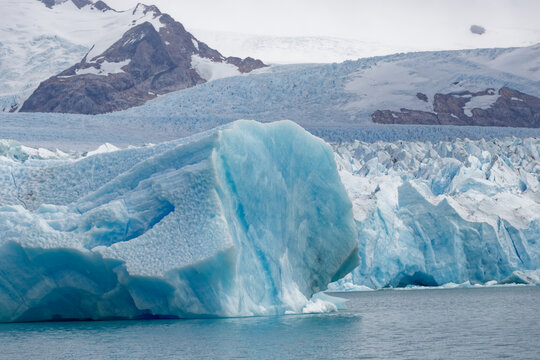 Massive cyan blue iceberg floating on cold lake waters near a melting glacier terminus against rugged snowy Patagonian mountain peaks.