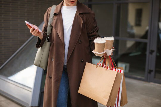 Close-up of a woman standing in a city street using her mobile phone while holding two takeaway coffees in a paper tray and three paper shopping bags