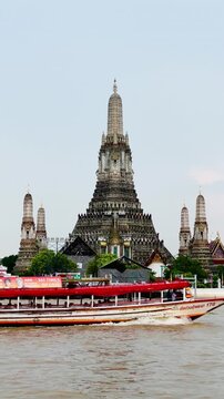 Wat Arun temple rises beside the Chao Phraya River in Bangkok, Thailand, as a long-tail boat cruises past during a peaceful evening with soft clouds and warm sunset light over the riverside.