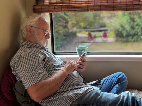 Side view of a senior man sitting by a window drinking a cup of coffee and using a mobile phone