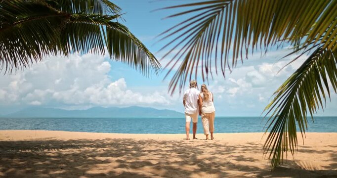 Senior couple walking hand in hand along sandy tropical beach, enjoying a relaxing vacation under palm trees with the clear blue ocean and distant mountains. Travel and relax on Samui Island, Thailand