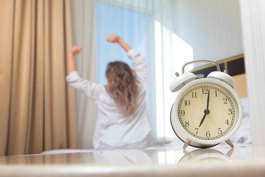 White alarm clock on glass table foreground. Blurred woman in shirt stretches on bed near window. Morning wake up routine. Back view