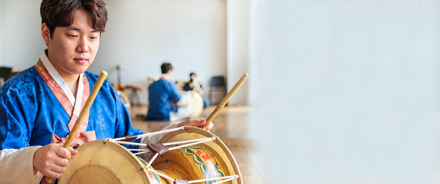 Young Korean drummer performing traditional percussion on a taut hide and lacquered wood drum, wearing a blue hanbok during Lunar New Year cultural rehearsal with open copy space and festive heritage