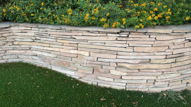 a curved retaining wall of stacked light stone slabs borders a lush lawn and is topped by flowering green bushes
