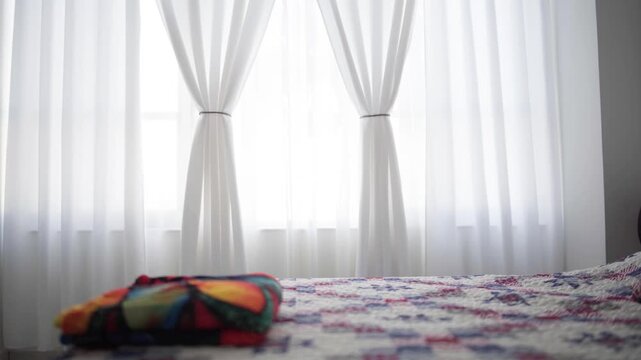 White window curtains hang in the bedroom. A folded colorful blanket on the bed. Domestic home comfort. Morning natural light illuminates the residential interior. Cozy bedding textile decor.