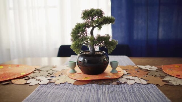 A potted artificial bonsai tree and egg cups stand on a wooden dining table. An autumn leaves table runner lies under the plant. Domestic home interior design setting. Cozy residential space.