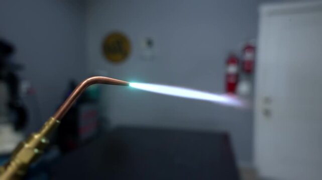 Handheld welding torch emits bright curved flame tracing curved copper tubing in dim workshop, warm sparks and glow highlighting metal texture