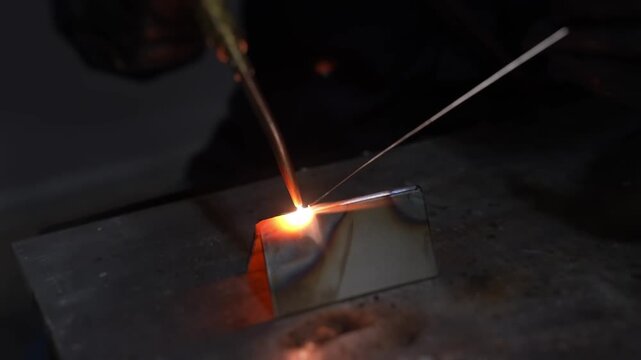Close up welding torch melting steel seam in dark workshop as bright orange arc sparks scatter over textured metal surface during manual precision work