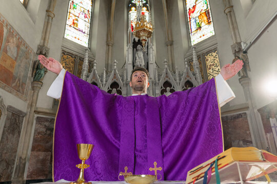 A parish priest stands with arms raised during a church ceremony. The setting is a catholic church where worship occurs.
