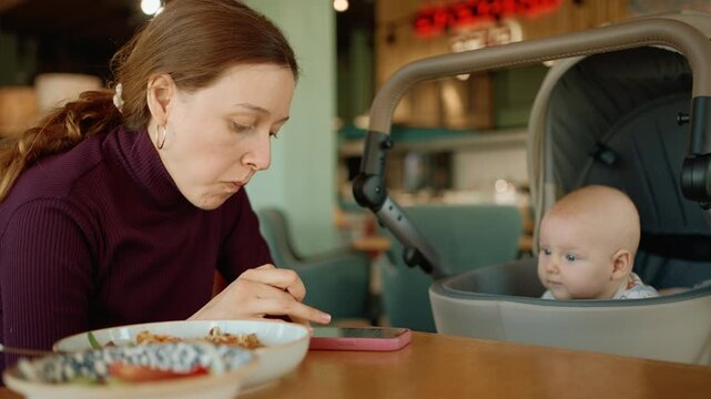 Young mother enjoying a meal in a cafe, simultaneously scrolling through her smartphone while her curious infant sits nearby in a stroller, looking around the public space