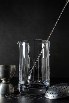 Empty cocktail beaker with jigger, julep strainer and bar spoon on dark surface 