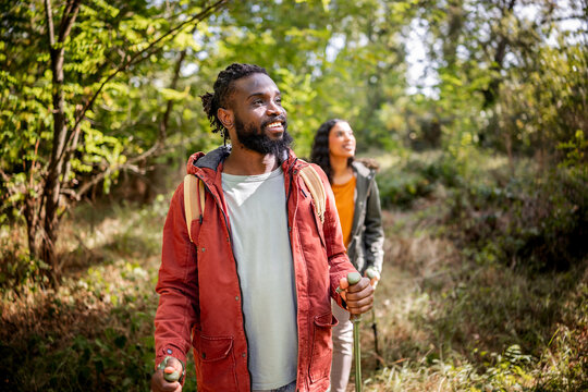 A joyful Black couple explores a vibrant forest, smiling and enjoying nature. The man, wearing a red jacket, holds hiking poles, while the woman, dressed in a green coat, looks up, appreciating the su