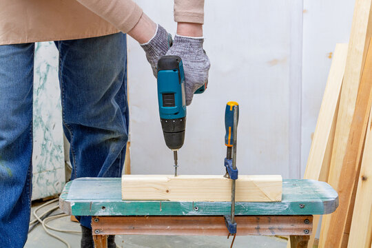 Person using a power drill to fasten a screw into wood on a workbench. DIY woodworking and home repair. Power tool use for a hands-on workshop project. Building and renovation